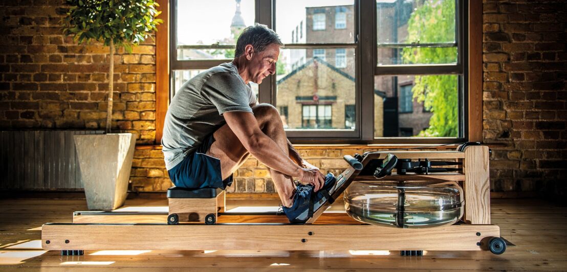 Ein Mann in Sportkleidung sitzt auf einem Rudergerät aus Holz, das mit einem echten Wassertank ausgestattet ist, im Hintergrund sieht man die dunklen Backsteine einer Loftwohnung