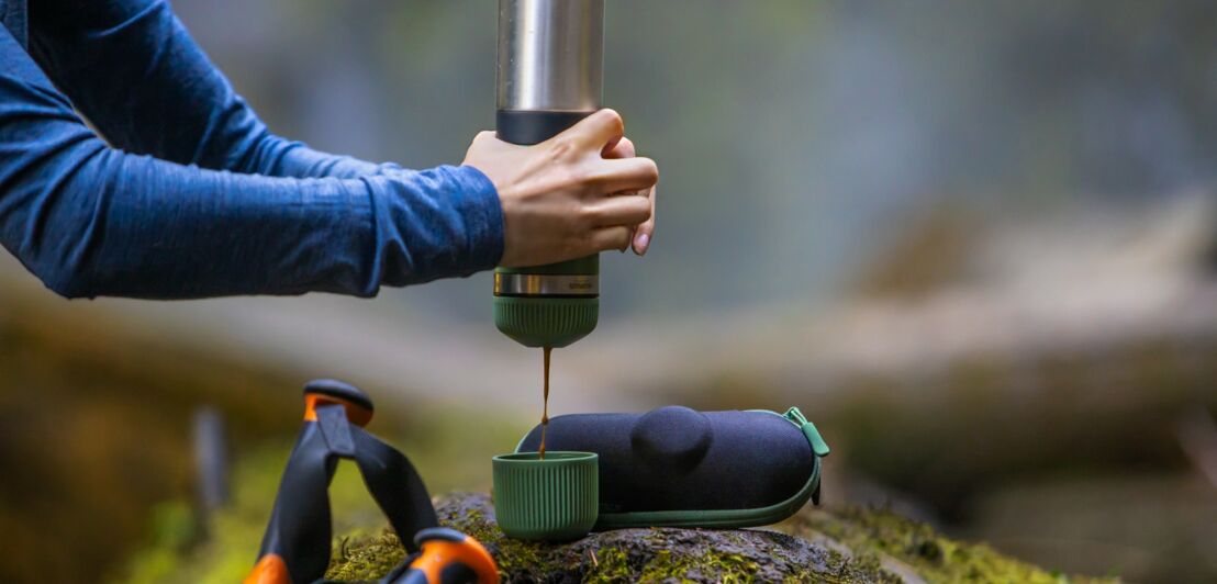 Eine Frau gießt sich bei einer Wanderung einen Espresso aus einer Thermoskanne in einen Becher.