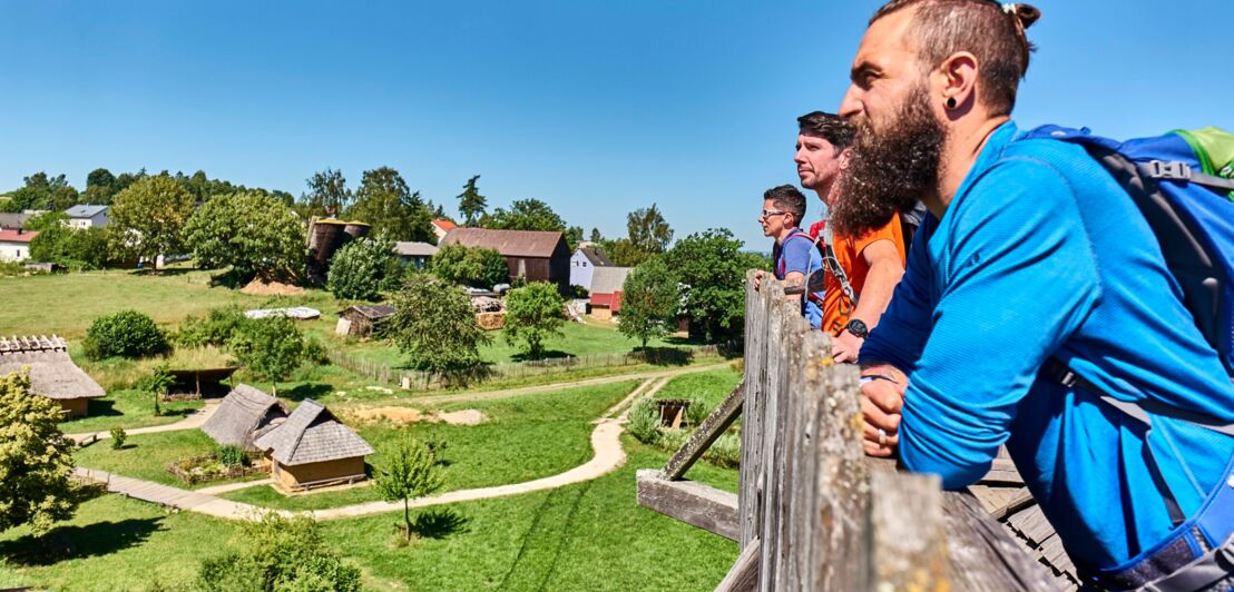 Wandernde stehen an einem Holzzaun von dem aus sie auf den Geschichtspark blicken