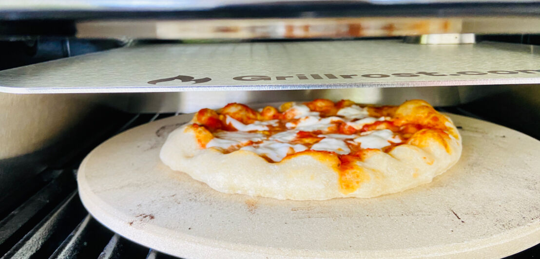 Pizza auf einem runden Stein der auf einem Rost liegt und ein Metallaufsatz darüber