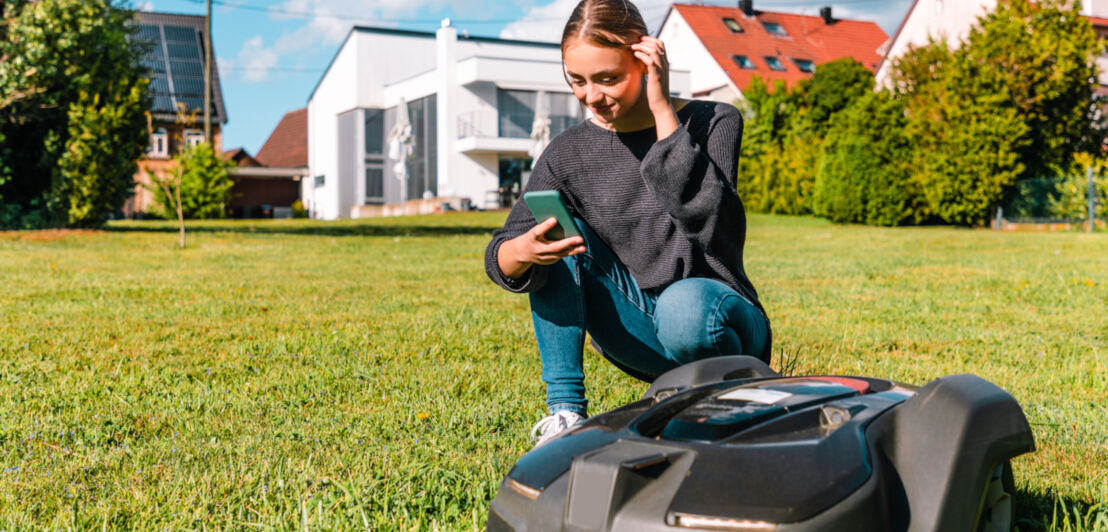 Eine Frau kniet mit dem Handy in der Hand vor einem Mähroboter