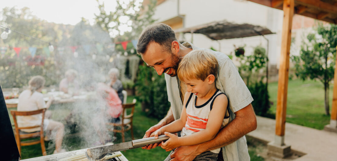 Ein Vater mit seinem Sohn auf dem Arm, der mit einer Grillzange einen Maiskolben wendet