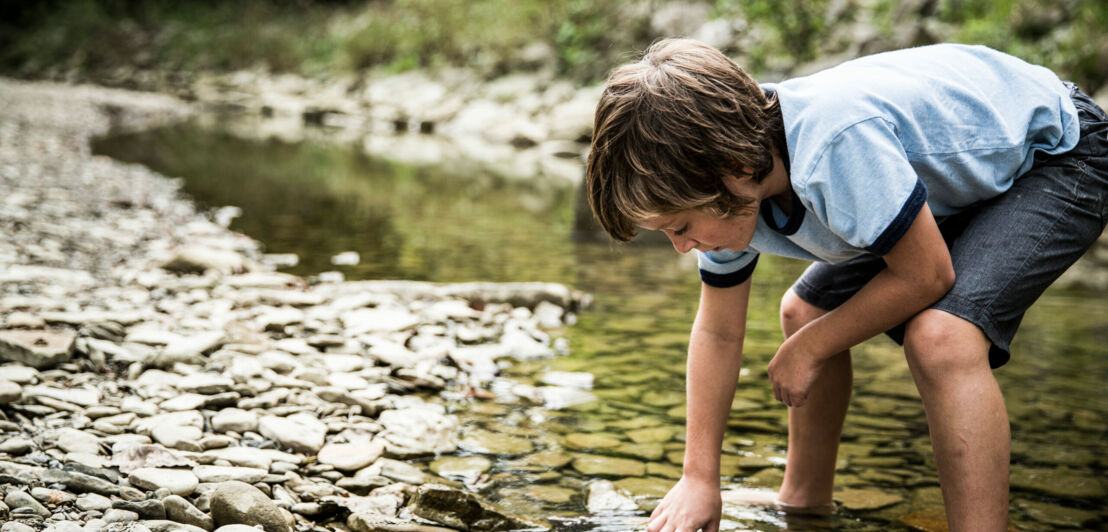 Ein Junge steht in einem Bachlauf und fasst ins Wasser