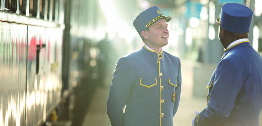 Ein Zug steht im Bahnhof, daneben zwei männliche Zugbegleiter in blauen Uniformen auf dem Bahnsteig