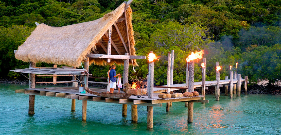 Ein überdachter Holzsteg im türkisblauen Wasser, darauf eine Familie am Lagerfeuer