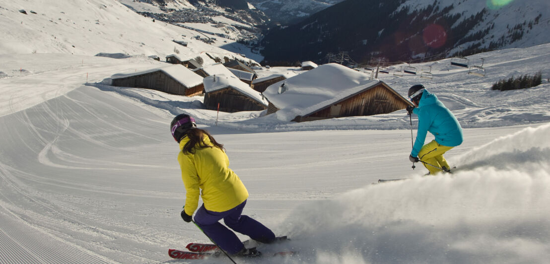 Zwei Skifahrer:innen fahren eine Abfahrt hinab.