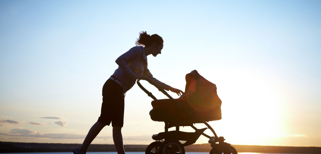 Frau als Silhouette im Gegenlicht streckt ihren Arm nach Kinderwagen aus, den sie neben einem Gewässer im Sonnenuntergang schiebt