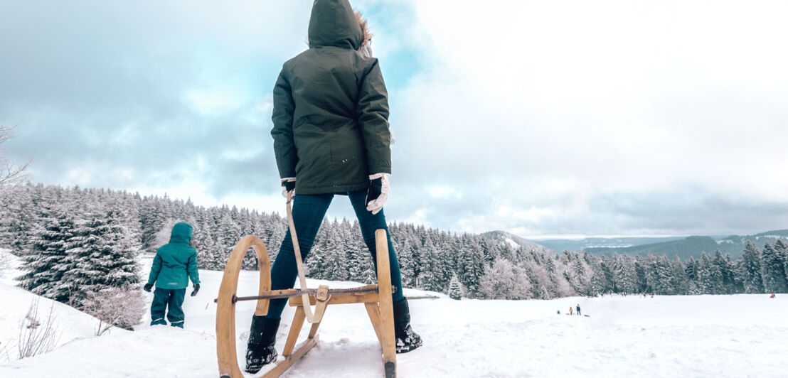 Zwei Kinder mit Schlitten auf einer Schneefläche, im Hintergrund Wälder und Berge.