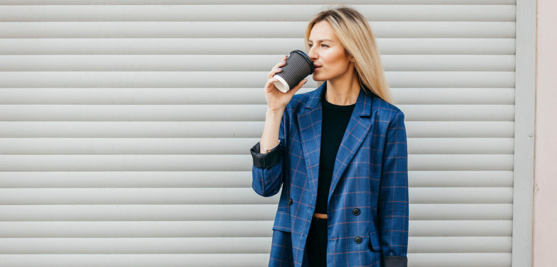 Eine Frau in einem blauen Blazer steht vor einer Garage und trinkt aus einem Becher