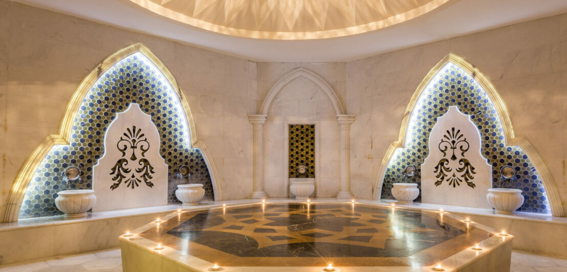 Elegantes, orientalischs Hamam mit goldfarbenem Nabelstein im Zentrum, darauf brennende Kerzen