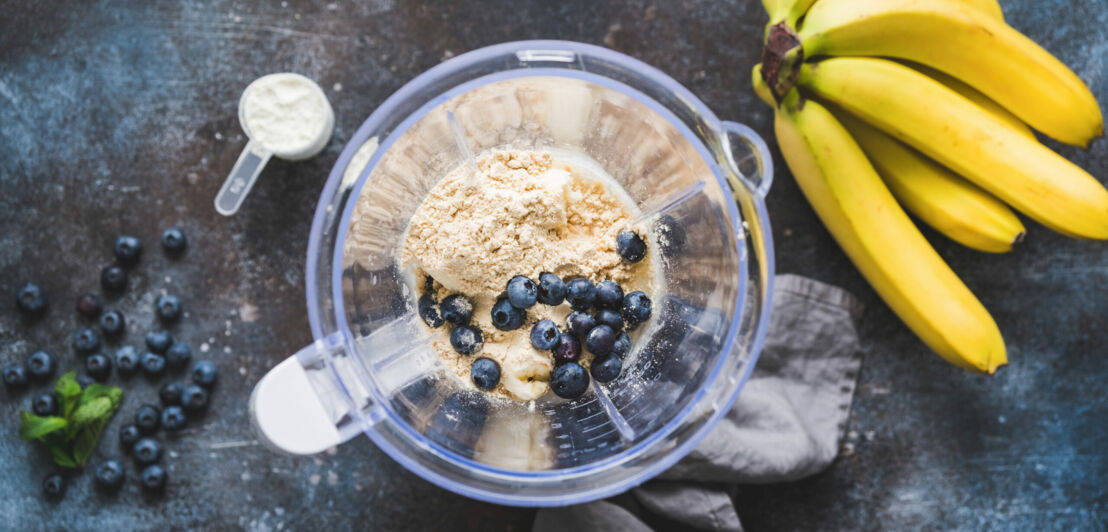 Mixer von oben, gefüllt mit Pulver und Beeren, daneben Bananen