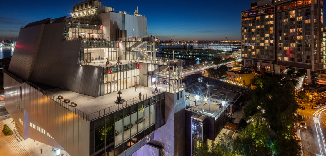 Beleuchteter, moderner Gebäudekomplex des Whitney Museum of Art bei Nacht aus der Luftperspektive, im Hintergrund der Hudson River