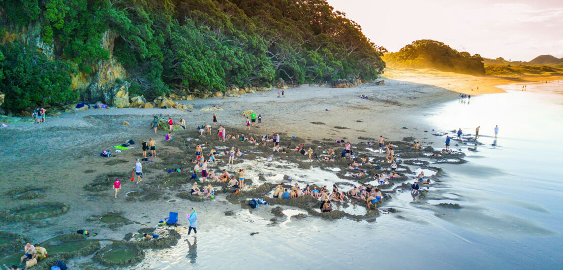 Ein Strand mit vielen sitzenden Menschen, im Hintergrund ein bewaldeter Felsen