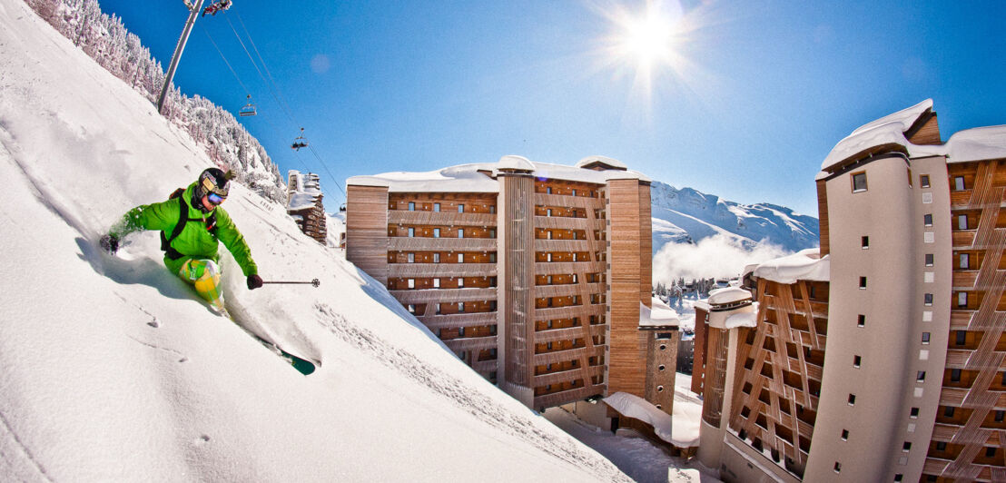 Eine Person mit Ski fährt einen steilen Hang hinab, im Hintergrund sind Häuser und Berge.