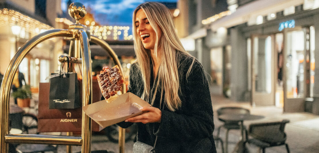 Eine lachende Frau mit einer frischen Waffel in der Hand, neben ihr steht ein Gepäckwagen mit Tüten