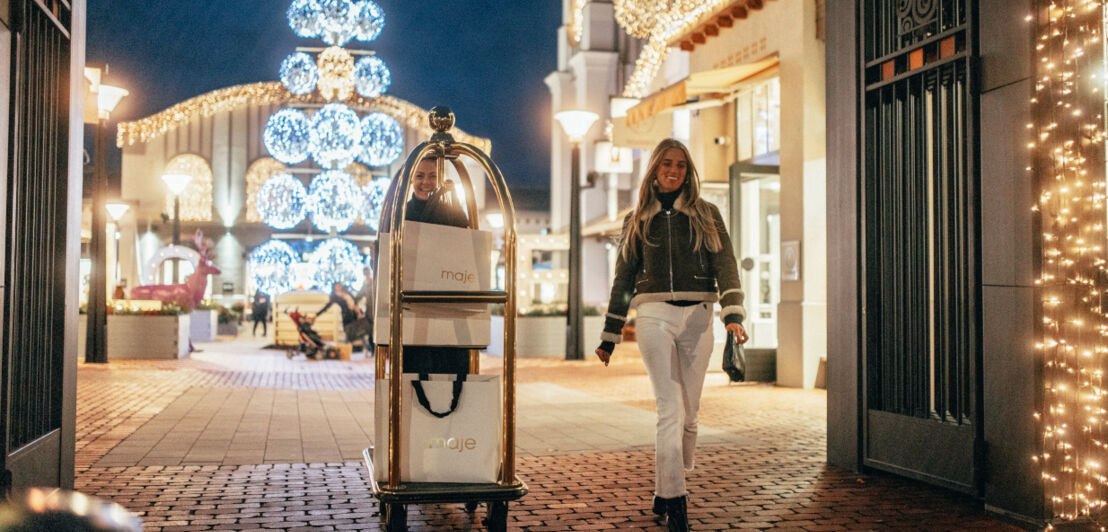 Eine Frau in weißer Jeans und Pilotenjacke läuft neben einer Concierge, die einen Trolley mit Einkaufstüten schiebt