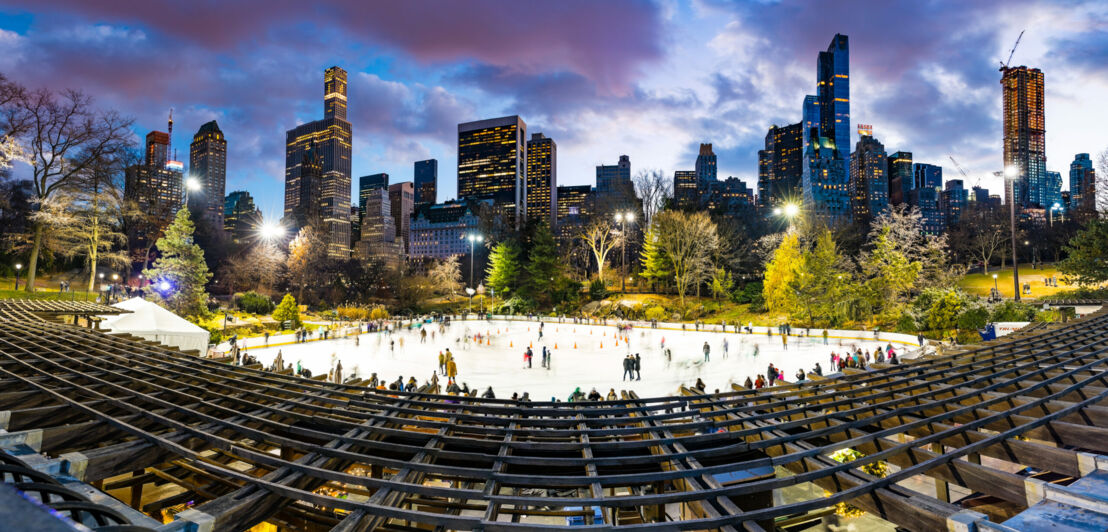 Weitwinkelaufnahme der beleuchteten und belebten Eisbahn im Central Park mit Skyline im Hintergrund in der Abenddämmerung