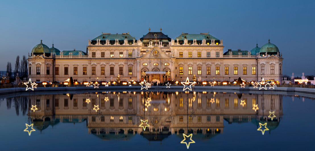 Das erleuchtete Wiener Barockschloss Belvedere mit Weihnachtsmarkt spiegelt sich in der Wasseroberfläche in der Abenddämmerung