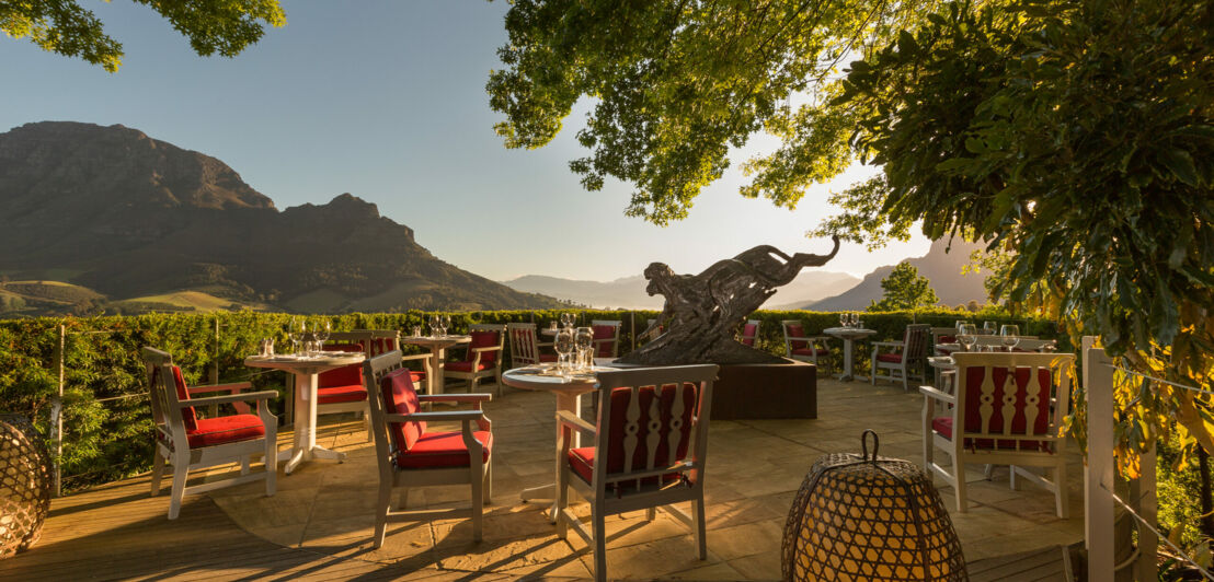 Elegante Terrasse mit weißen Tischen und roten Polsterstühlen mit Blick auf eine gebirgige Landschaft in der Abendsonne