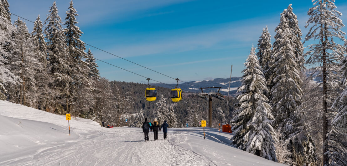 Drei Personen gehen eine Skipiste hinab, über ihnen fahren gelbe Gondeln hinweg