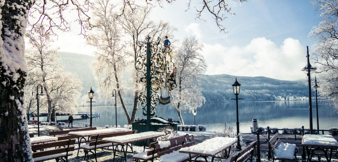 Ein mit Schnee bedeckter Biergarten vor einem See, im Hintergrund Berge