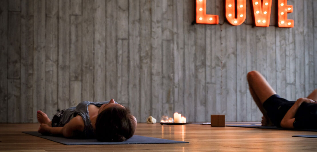 Zwei Personen liegen in einem gemütlichen Yoga-Raum auf ihren Matten, auf dem Boden stehen Kerzen und an der Wand leuchten die Lettern LOVE