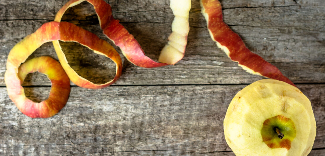 Ein geschälter Apfel und seine Rinde, die von oben fotografiert auf einem Holztisch liegen