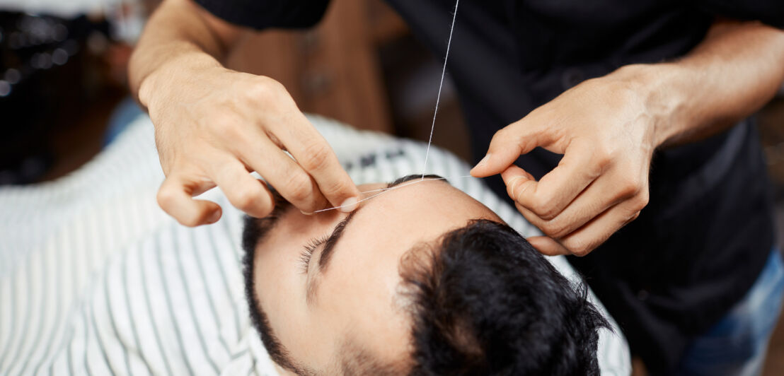 Ein Mann modelliert die Augenbrauen seines Klienten, der in einem Friseurstuhl liegt