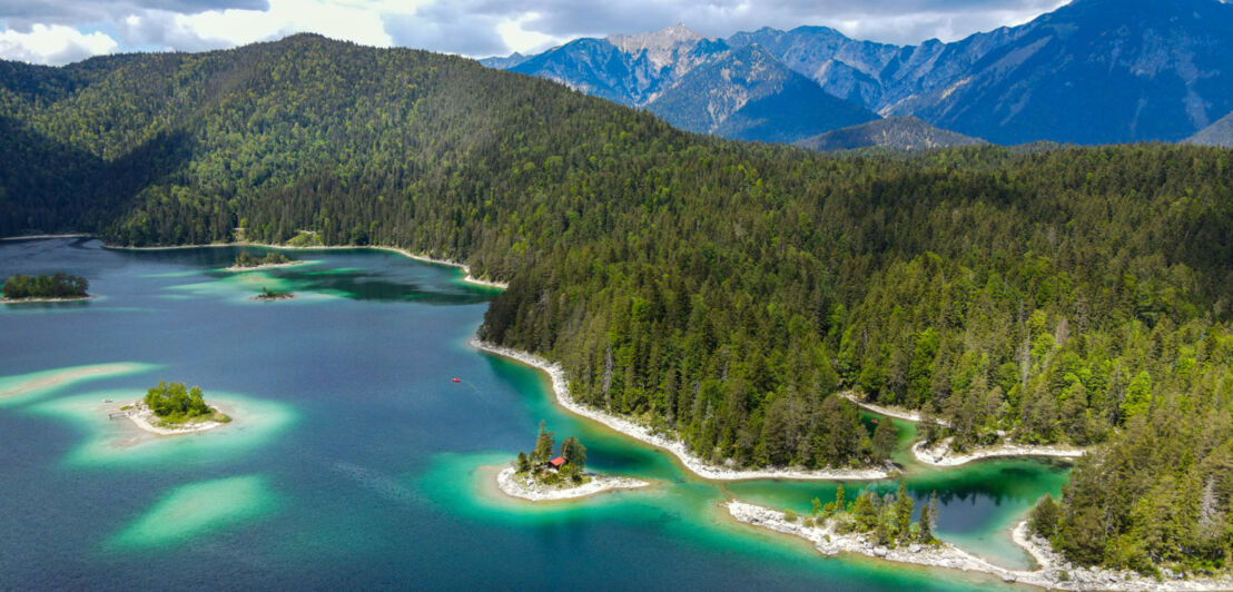 Aufsicht auf einen See mit türkisblauem Wasser und vorgelagerten Inseln inmitten üppiger Waldlandschaft, im Hintergrund Bergpanorama