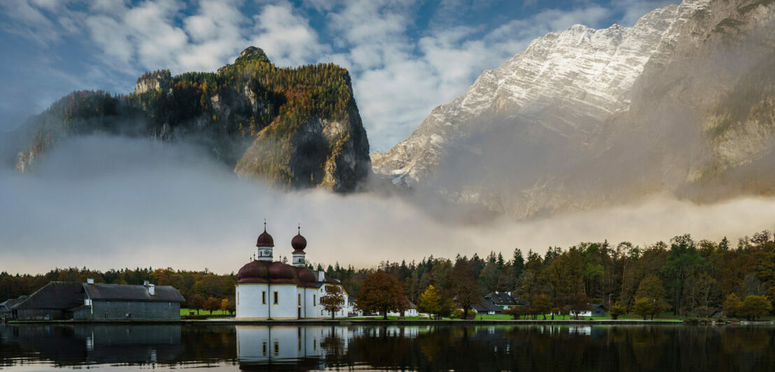 Eine weiße Kapelle mit roten Zwiebeltürmen in der herbstlichen Landschaft am Ufer eines Sees vor zwei riesigen Felswänden