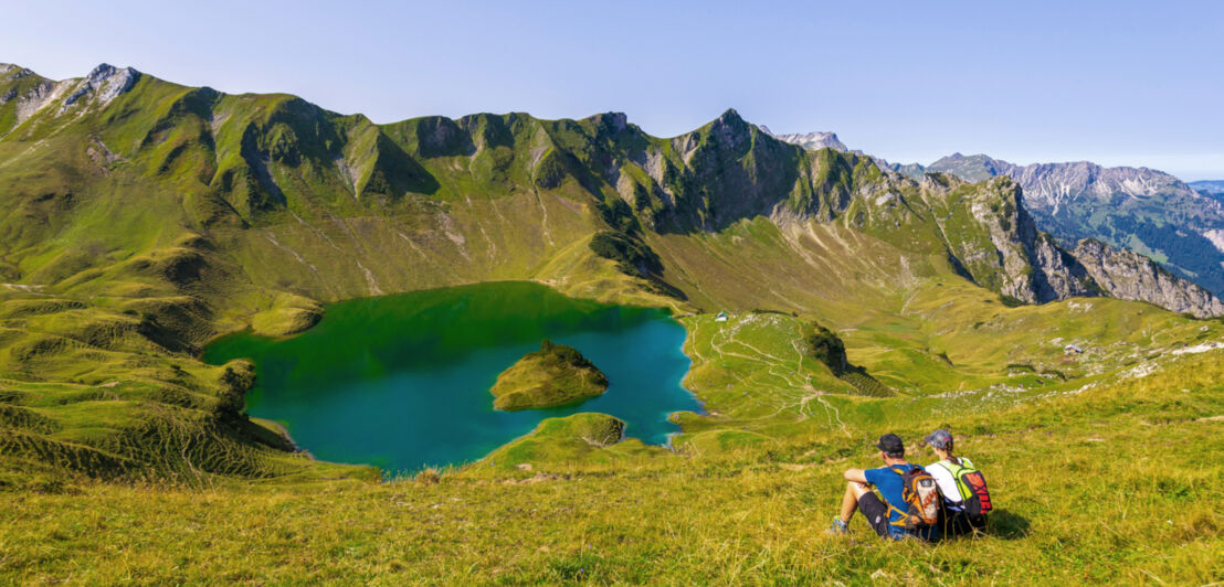 Zwei Wandernde sitzen mit Rucksäcken auf der Wiese vor einem Bergsee