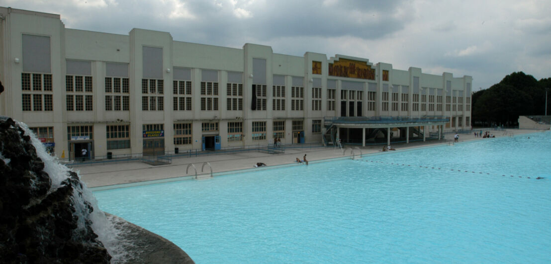 Ein großes blaues Schwimmbecken vor dem Hintergrund eines genauso großen Gebäudes im Art-déco-Stil