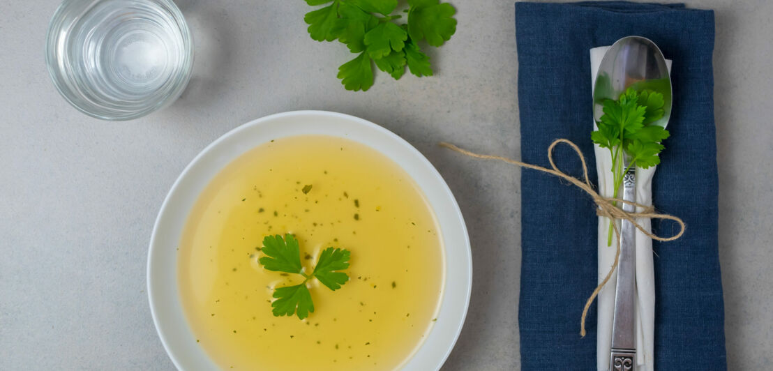 Klare Suppe in einem weißen Teller mit Petersilie, daneben ein Löffel und ein Wasserglas