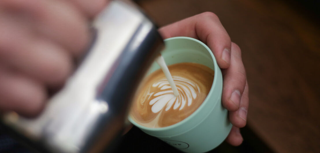 Eine Hand gießt aufgeschäumte Milch in einen Plastikbecher mit Kaffee