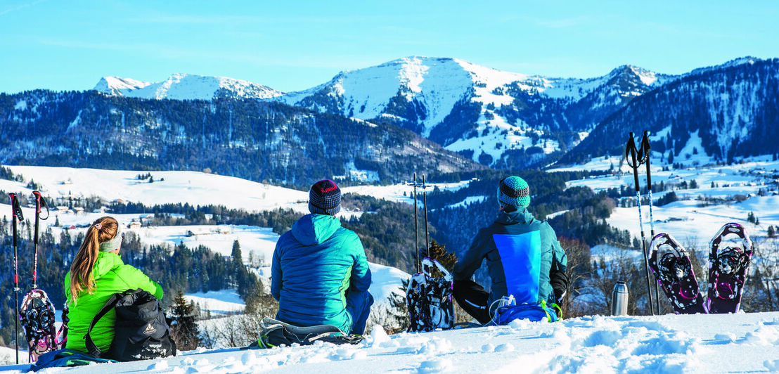 Drei Menschen in Winterkleidung sitzen im Schnee und blicken in die Ferne