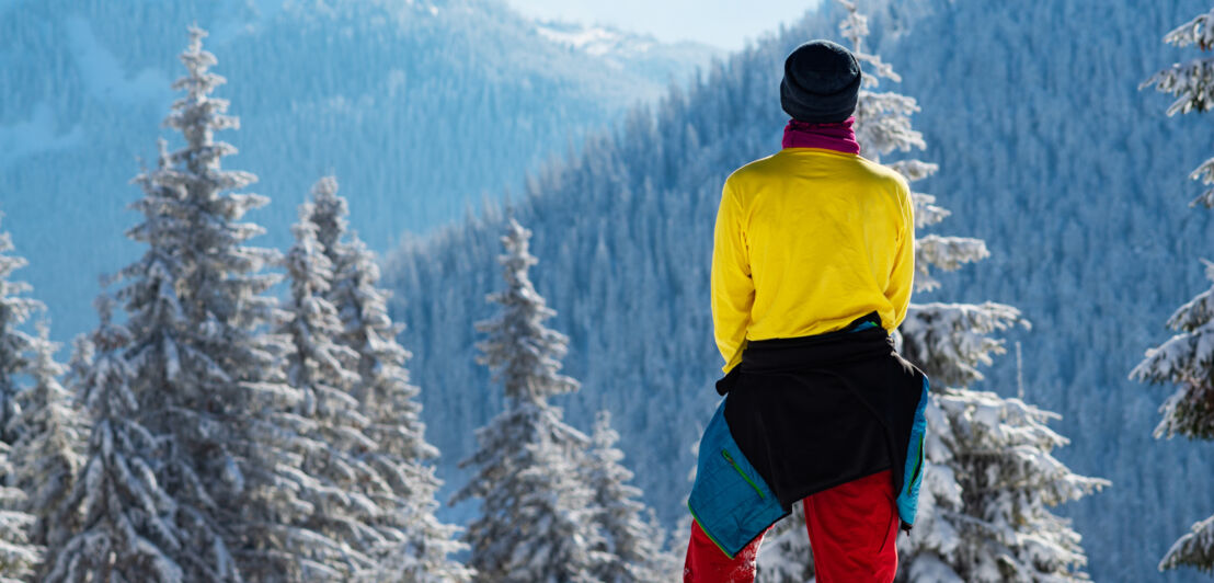 Eine Person mit Mütze, Schal und um die Hüfte gebundener Jacke blickt auf eine Berglandschaft