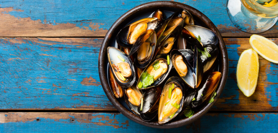 Frische, marinierte Miesmuscheln in einer Schale auf blau gestrichenem Holzboden, daneben zwei Stückchen Zitrone und ein Glas Weißwein