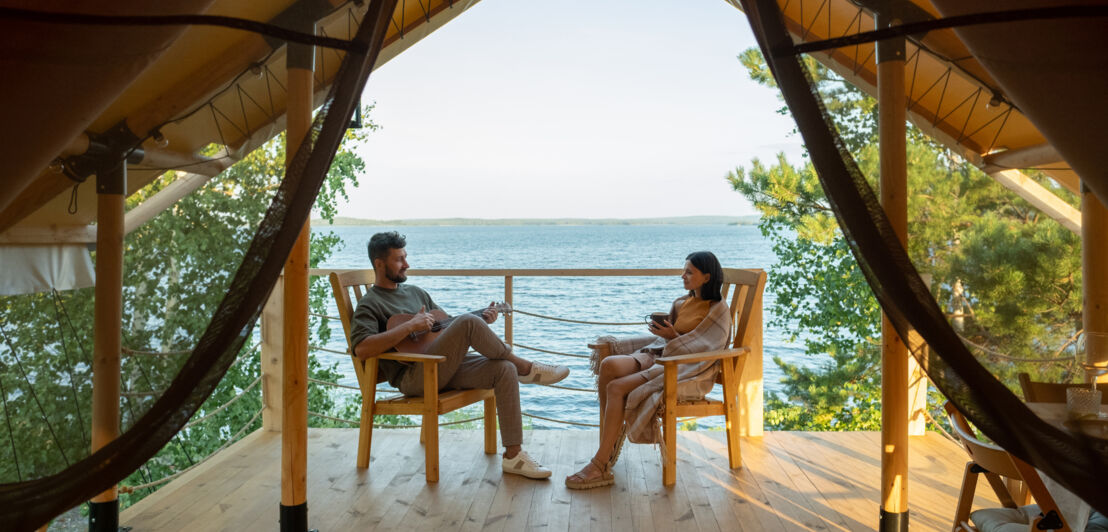 Pärchen in Holzstühlen sitzt vor einer Glampingunterkunft mit Blick auf das Meer.