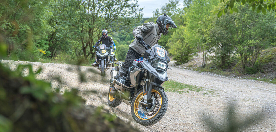 Zwei Reiseenduros der Marke BMW fahren hintereinander über eine bewaldete Schotterstrecke.