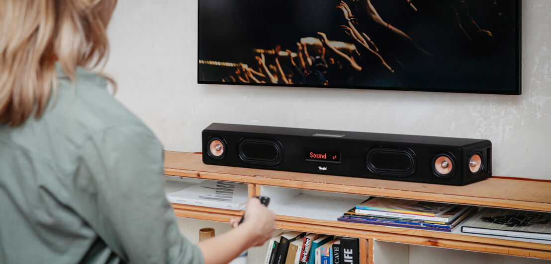 Eine Frau bedient mit einer Fernbedienung eine Soundbar unterhalb eines TV-Gerätes