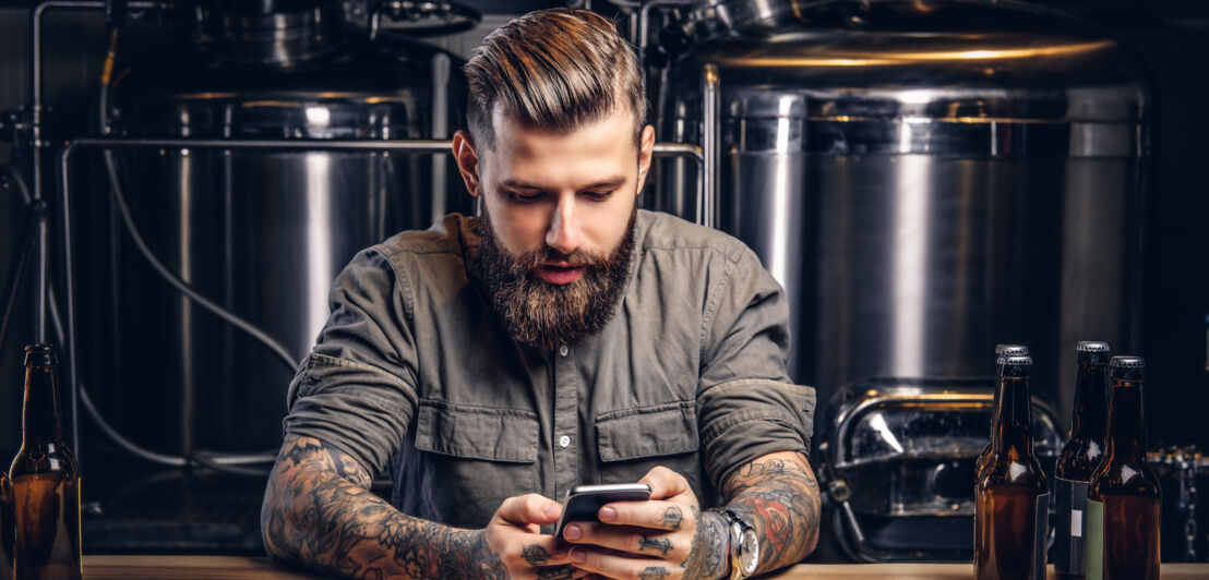 Ein Mann sitzt mit einem Smartphone an einem Tisch, auf dem Bierflaschen stehen, im Hintergrund Bierkessel