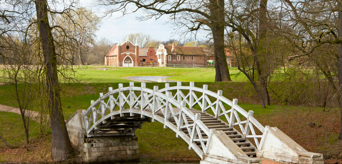 Eine kleine Brücke über einem Flusslauf, dahinter eine Wiese und ein großes Anwesen