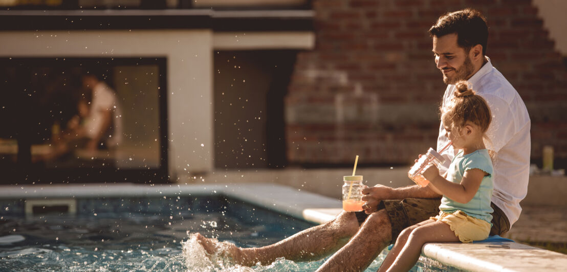 Ein Mann und seine Tochter planschen mit den Beinen im Wasser am Rand eines Pools und trinken Limonade