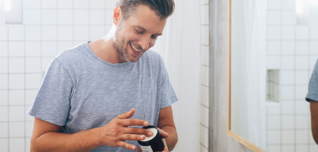 Ein lachender Mann steht im Bad und hat einen Bottich Creme in der Hand.