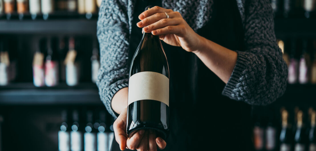 Eine Frau hält eine Flasche Wein in der Hand und präsentiert sie in die Kamera.