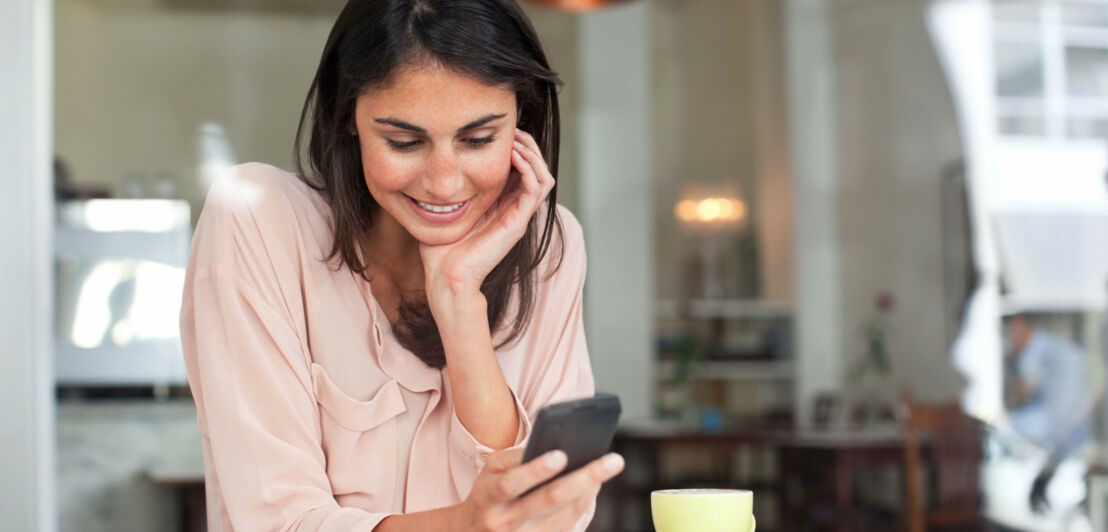 Eine junge Frau an einem Tisch in einem Café schaut lächelnd auf ihr Smartphone