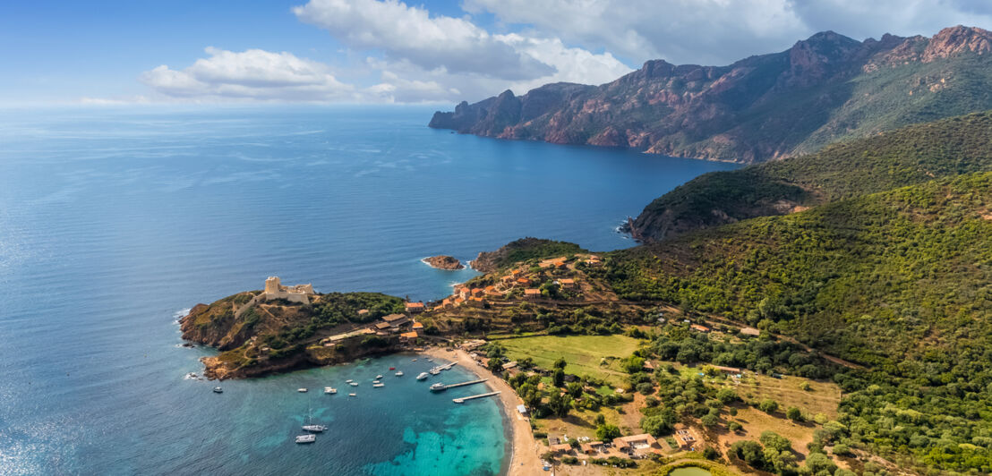 Blick über eine Bucht auf Korsika mit türkisem Wasser, ein paar Booten und einer Gebirgskette im Hintergrund.