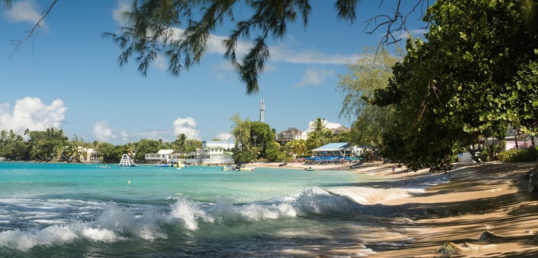 Ein Strandabschnitt mit türkisblauem Wasser und Bäumen, im Hintergrund einige Beach-Häuser.