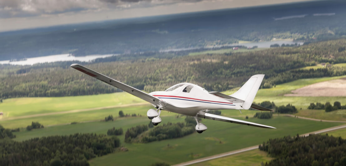 Ein Ultraleichtflugzeug, das über eine flache Landschaft mit Wäldern, Feldern und Fluss fliegt