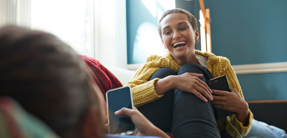 Zwei junge, vertraute Menschen mit Smartphones in den Händen genießen lachend die gemeinsame Zeit auf einem Sofa